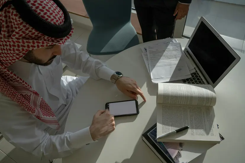 A man in a ghutra working through papers and a laptop at an office table