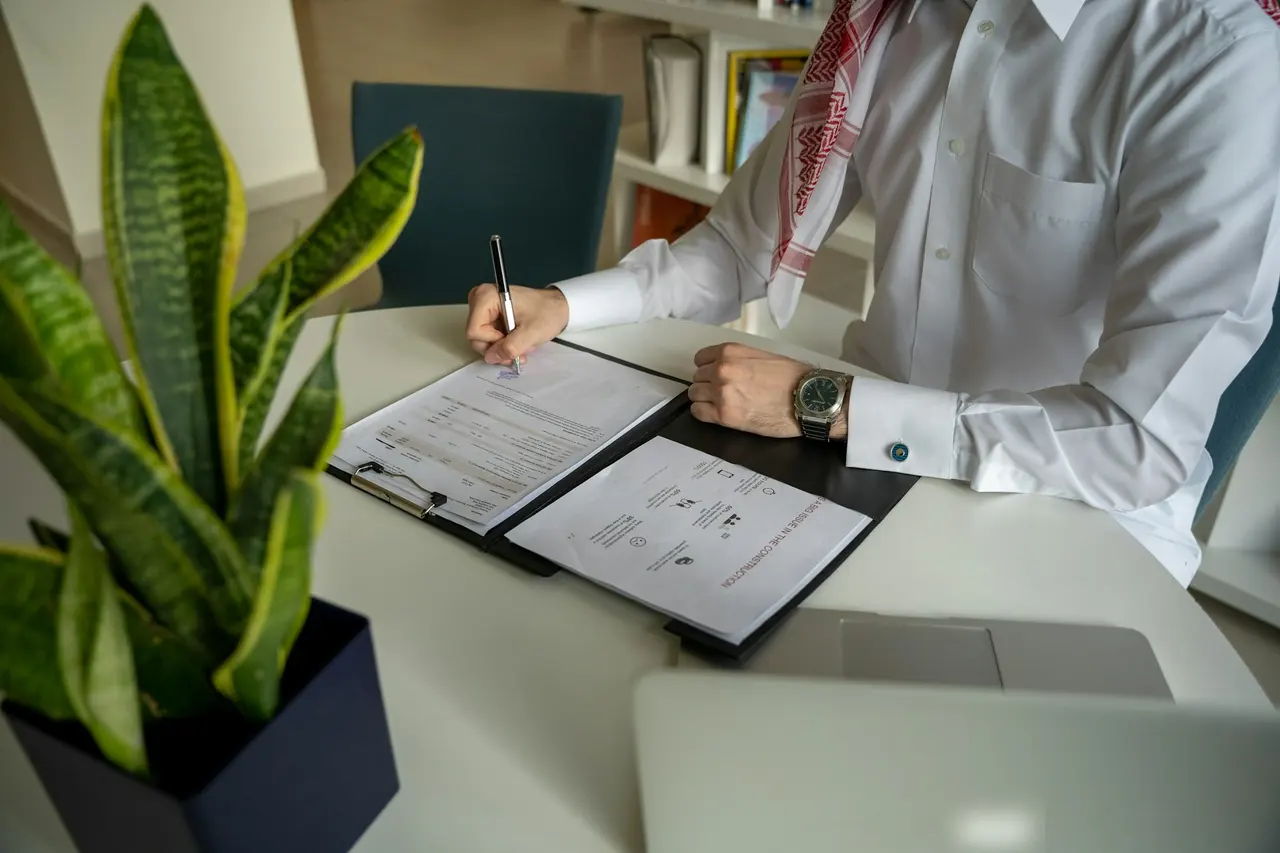 A man in a ghutra writing notes in a folder at an office desk in Doha