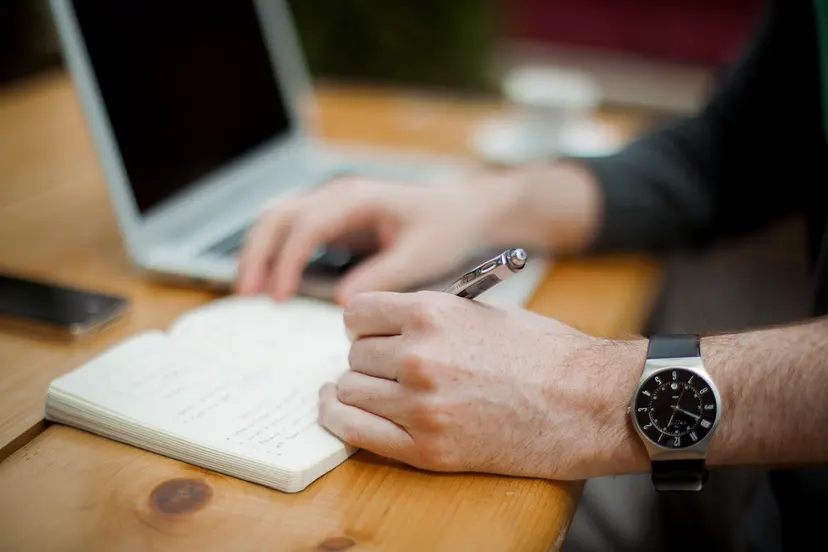 A hand writing in a notebook beside a laptop