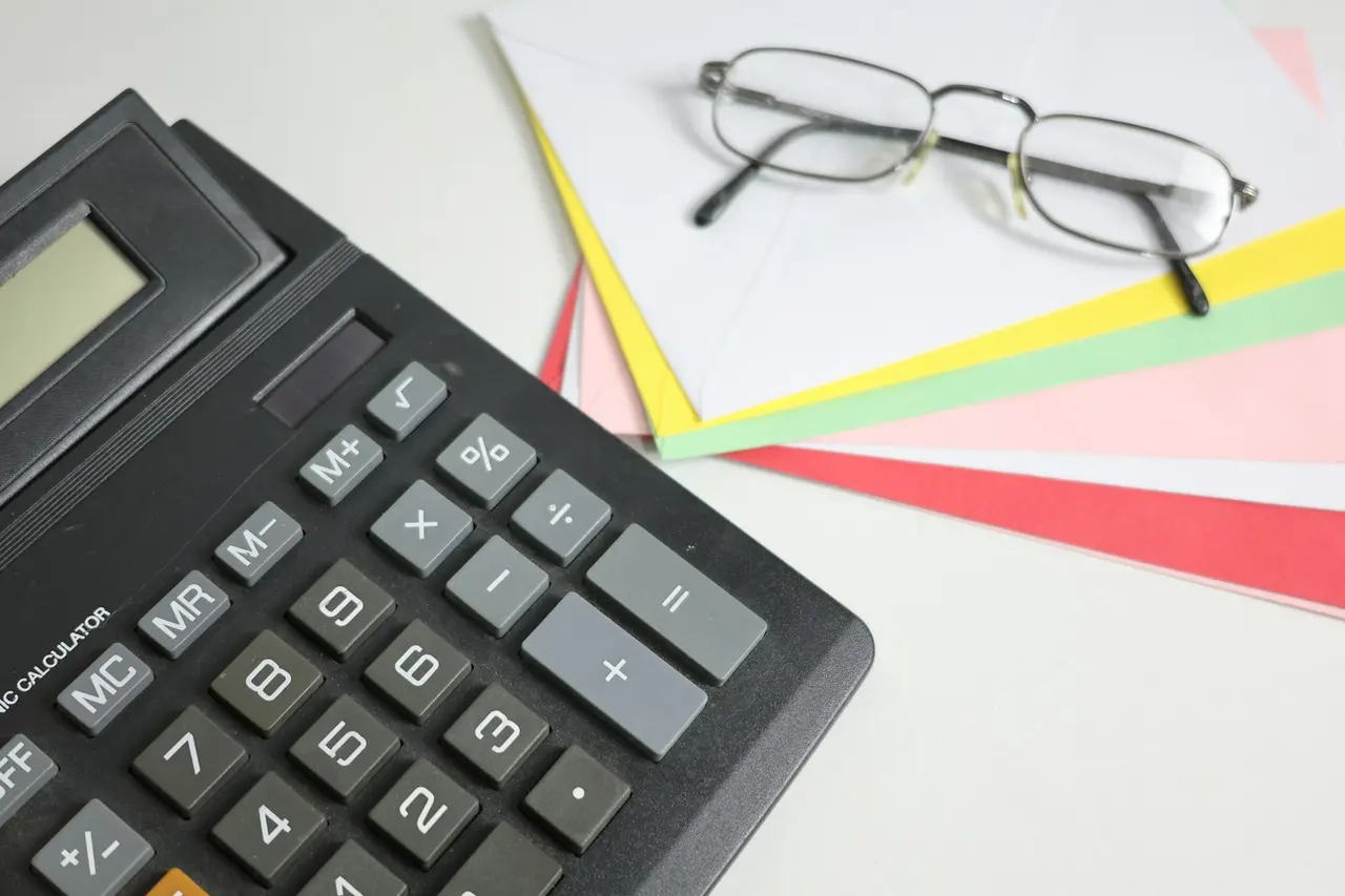 A desk calculator beside reading glasses and coloured envelopes
