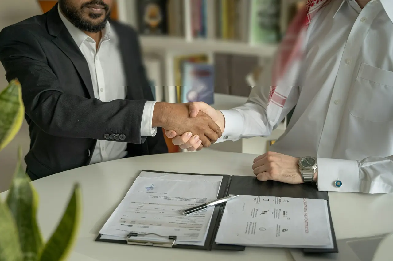 Two businessmen shaking hands across a desk with documents