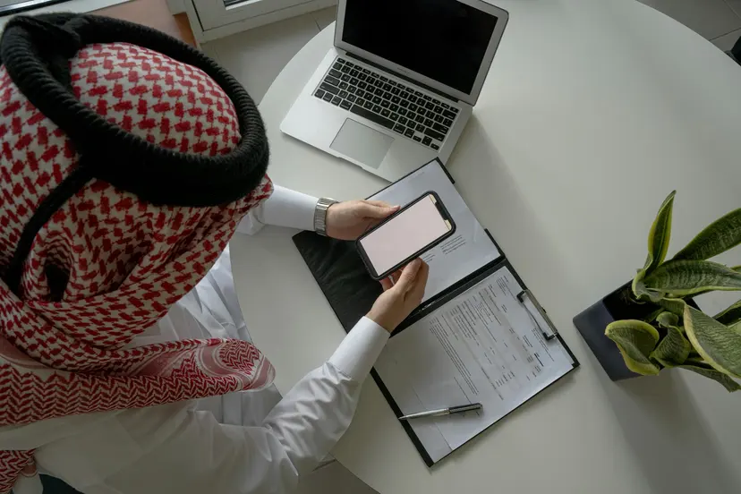 A man in a ghutra working at a desk with a laptop, phone and clipboard